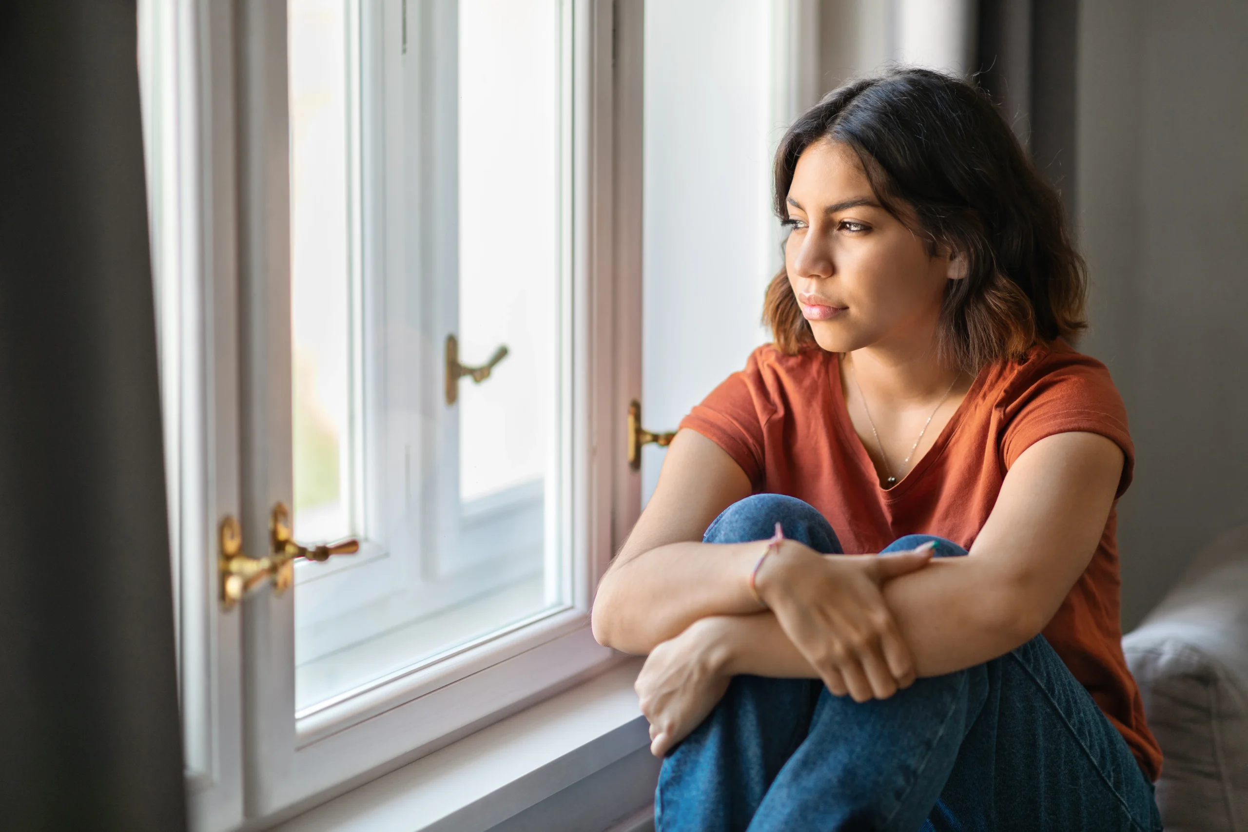 Woman sitting by a window looking pensive, reflecting the extreme hardship a qualifying relative may face