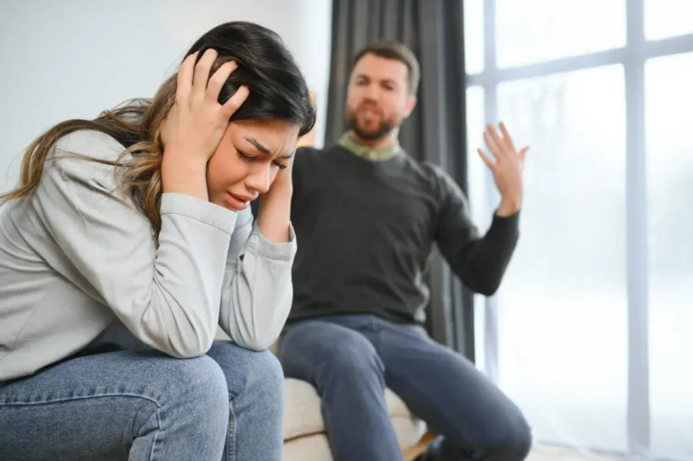 Distressed woman holding her head while a man gestures behind her — representing the emotional impact of domestic abuse documented in a VAWA psychological evaluation